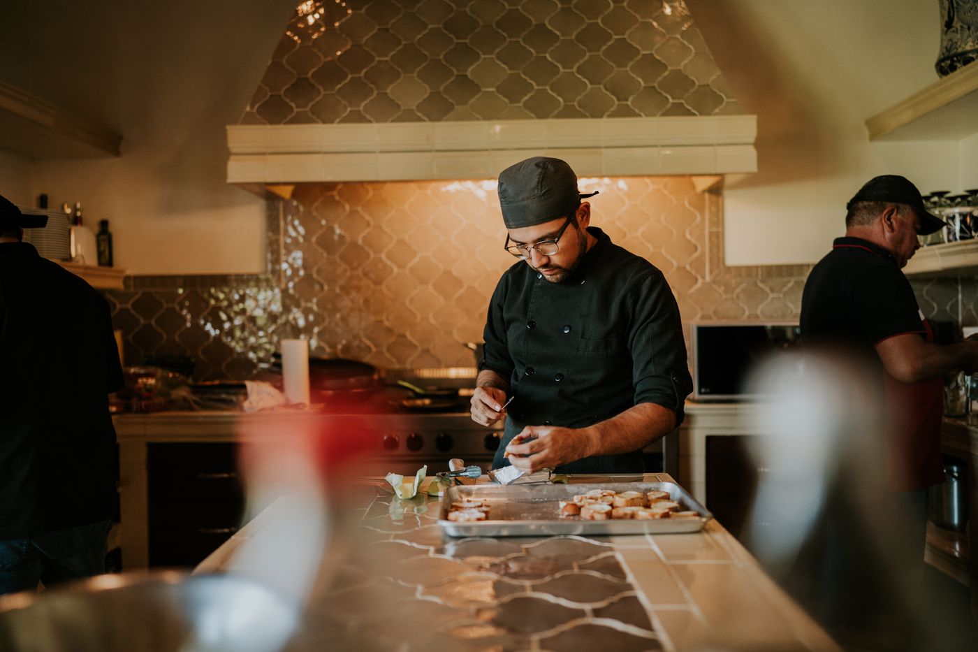 A chef in a dark jacket and cap preparing food at a steel counter in a working restaurant kitchen