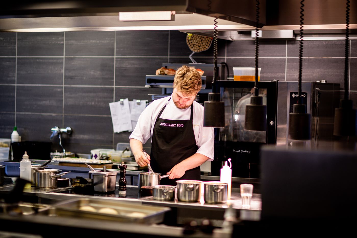 A chef in a black apron preparing food at the pass of a modern restaurant kitchen