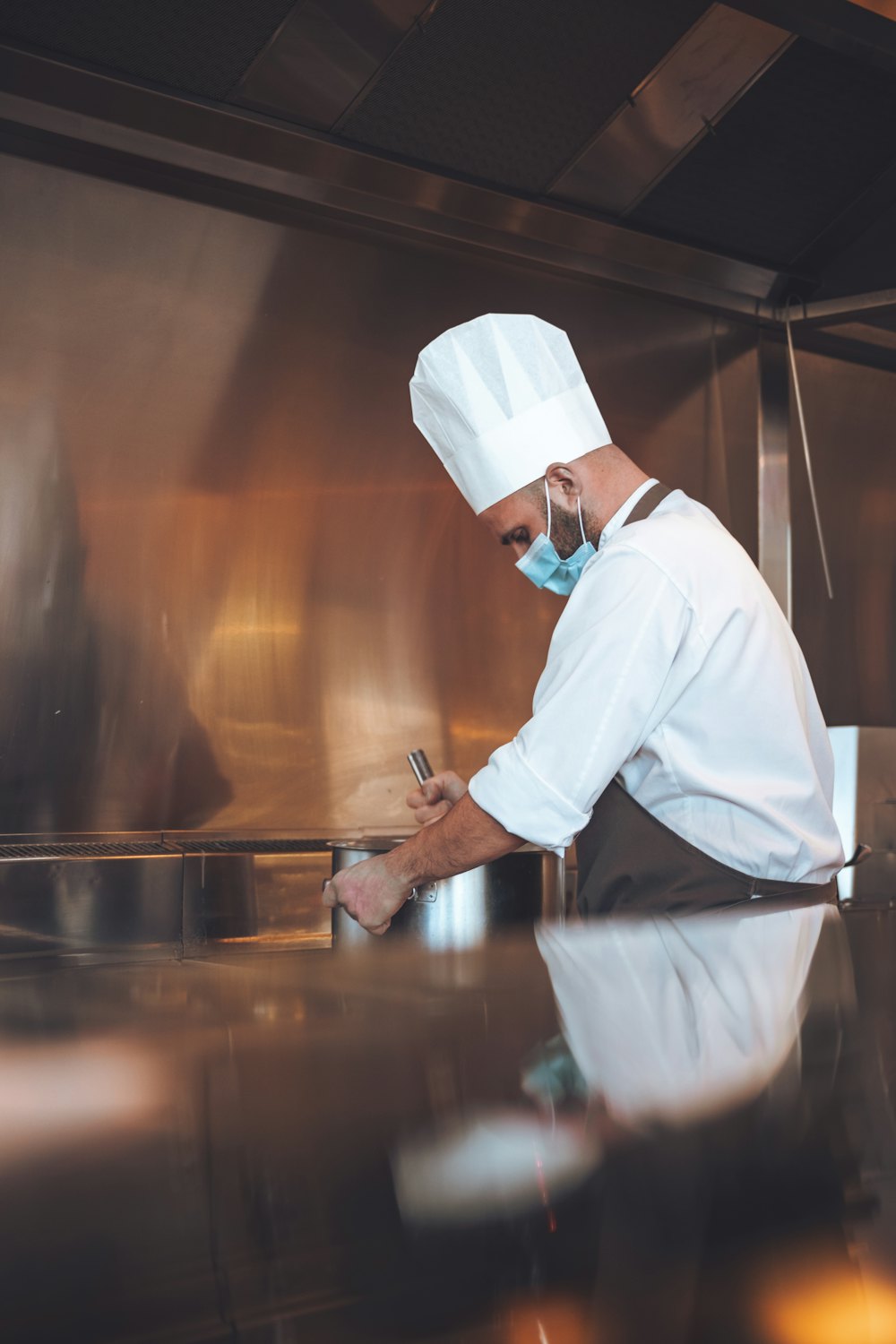 A chef in a toque and face mask making notes at a stainless-steel kitchen counter