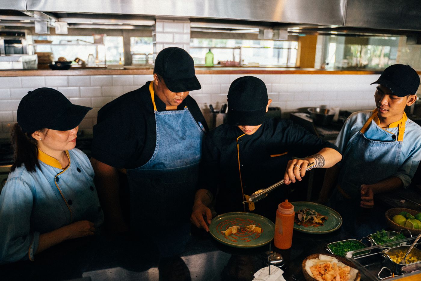 A kitchen team in caps and aprons gathered around a chef demonstrating at the pass
