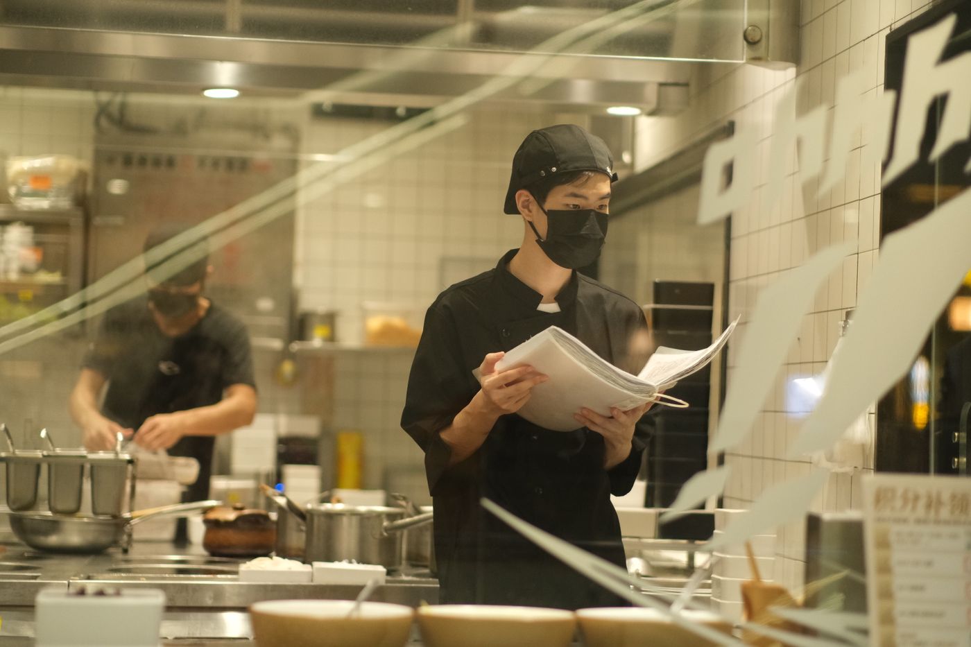 A masked cook in a cap reviewing a printed checklist in a working commercial kitchen